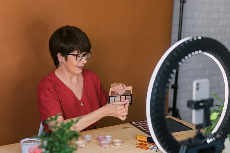 Foto blogger de belleza femenina de mediana edad o influencer con producto de maquillaje grabando video en la habitación en casa usando lámpara de anillo y teléfono inteligente