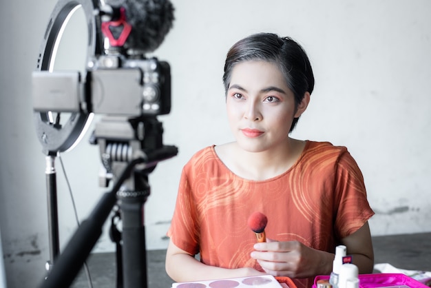 Blogger de belleza de moda hermosa asiática mira en una cámara digital. revisa productos de belleza para videoblog, hablando sobre cosméticos sosteniendo una paleta de maquillaje mientras graba su video.