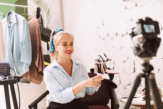 Blogger de belleza sonriente sosteniendo zapatos en el talón en las manos mientras graba felizmente un nuevo video de moda para vlog en cámara