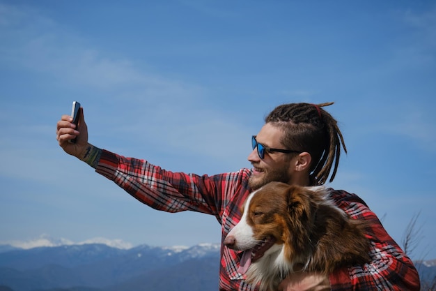 Blogger caucásico con gafas de sol se sienta en la cima de la montaña y se toma una selfie con un pastor australiano