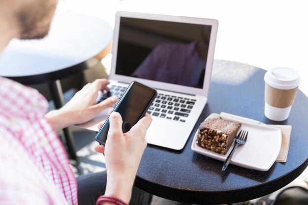 Blogger de comida que usa un teléfono inteligente y una computadora portátil mientras come pasteles en el café