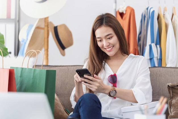 Blogger de diseñador de moda de hermosa mujer asiática sentada en la tienda de ropa y recibir el pedido del cliente con el teléfono inteligente. Influenciador en línea de negocios en concepto de redes sociales.