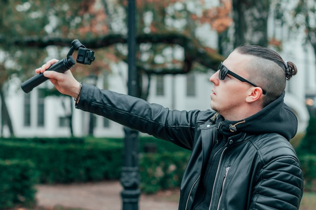 Blogger em óculos de sol fazendo selfie ou streaming de vídeo no parque outono usando câmera de ação com estabilizador de câmera gimbal.