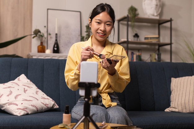 Blogger femenina asiática haciendo maquillaje filmando en el teléfono celular en casa