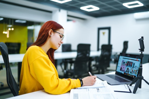 Blogger femenina con laptop grabando video mientras está sentada en la oficina y hablando de negocios de inicio