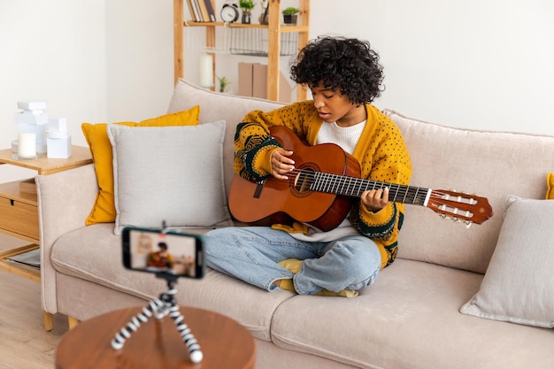 Blogger guitarrista Chica afroamericana bloguera tocando la guitarra hablando con cámara web grabando vlog Mujer influyente en las redes sociales transmitiendo en casa en interiores Creador de contenido musical tutorial de transmisión