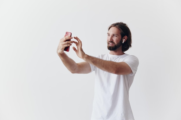 Un blogger masculino grabándose en video en su teléfono y chateando con personas en línea con una sonrisa en una camiseta blanca contra una pared blanca