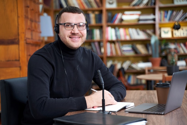 Blogger masculino que transmite desde la biblioteca usando una computadora portátil y un micrófono