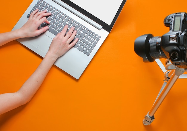 Foto blogger mujer escribiendo en una computadora portátil, blogueando con una cámara con un trípode sobre fondo naranja. tecnoblogging. revisión de la computadora portátil. vista superior