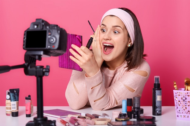 Blogger de mujer o belleza con pincel de sombra de ojos, espejo y cámara, sentada a la mesa, rodeada de cosméticos, grabando maquillaje