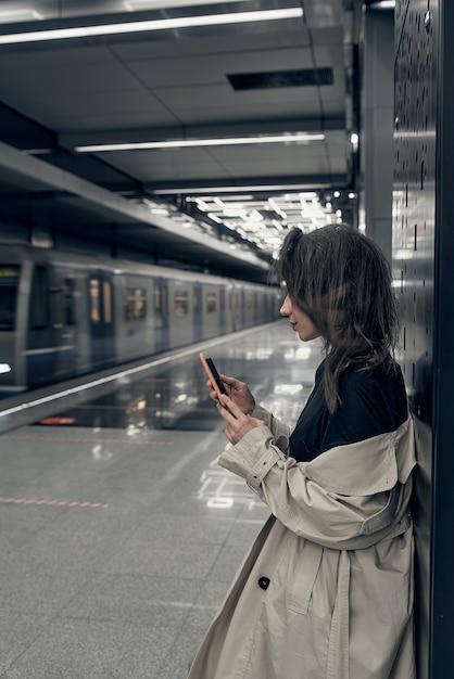 Bloggerin in einem beigen Trenchcoat in der U-Bahn, die auf einen Zug auf dem Bahnsteig wartet, benutzt ein Smartphone.