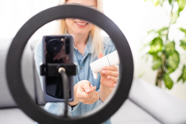 bloggerin nimmt video auf smartphone auf und beleuchtet sich mit ringlampe am tisch im hellen raum.