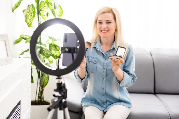 bloggerin nimmt video auf smartphone auf und beleuchtet sich mit ringlampe am tisch im hellen raum.