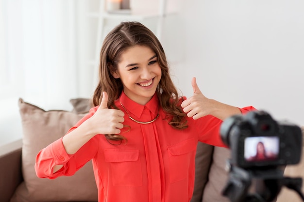 Blogging, tecnología, videoblog, medios de comunicación y concepto de personas: mujer sonriente feliz o bloguera con cámara grabando video y mostrando los pulgares hacia arriba en casa