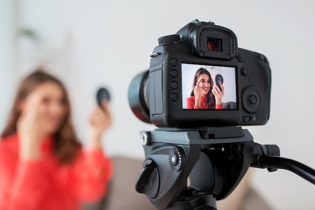 blogs, tecnología, videoblog y concepto de personas: mujer sonriente feliz o bloguera de belleza con lápiz de cejas, espejo y cámara grabando video tutorial de maquillaje en casa