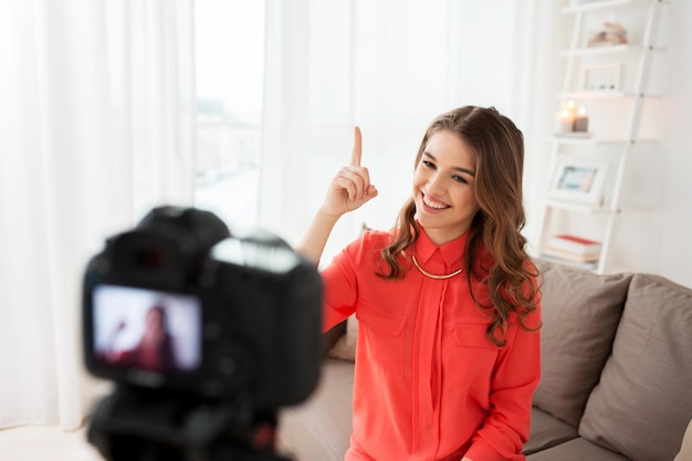 blogs, tecnología, videoblog, medios de comunicación y concepto de personas: mujer sonriente feliz o blogger con cámara grabando vídeo en casa y apuntando con el dedo hacia arriba