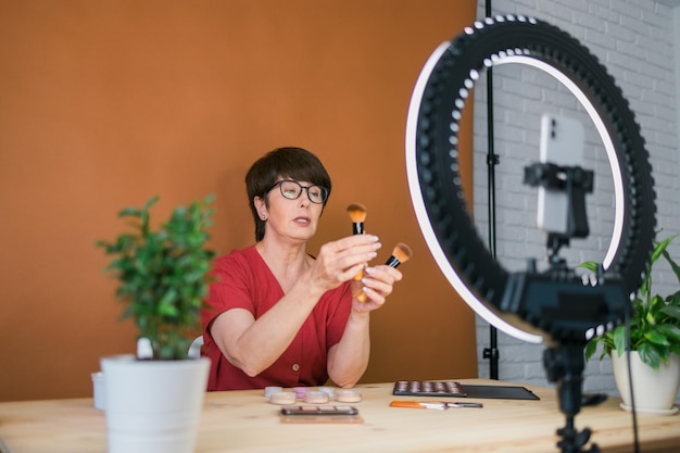 Blogueira de beleza feminina de meia-idade ou influenciadora com produto de maquiagem gravando vídeo no quarto em casa usando lâmpada de anel e smartphone