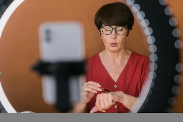 Blogueira de beleza feminina de meia-idade ou influenciadora com produto de maquiagem gravando vídeo no quarto em casa usando lâmpada de anel e smartphone