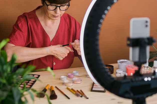 Blogueira de beleza feminina de meia-idade ou influenciadora com produto de maquiagem gravando vídeo no quarto em casa usando lâmpada de anel e smartphone