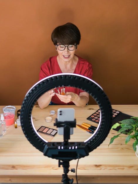 Blogueira de beleza feminina de meia-idade ou influenciadora com produto de maquiagem gravando vídeo no quarto em casa usando lâmpada de anel e smartphone