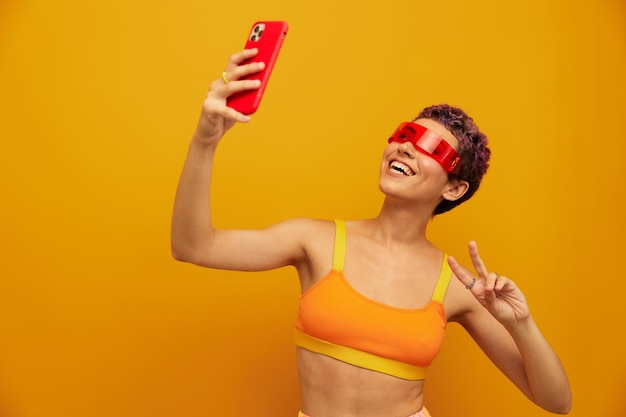 Blogueira de mulher sorrindo em óculos milenares incomuns tirando selfies em seu telefone em roupas esportivas contra um espaço livre de estúdio laranja