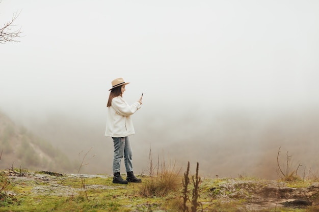 Blogueira elegante em uma colina fotografando um destino rochoso, uma floresta de pinheiros e colinas nebulosas.