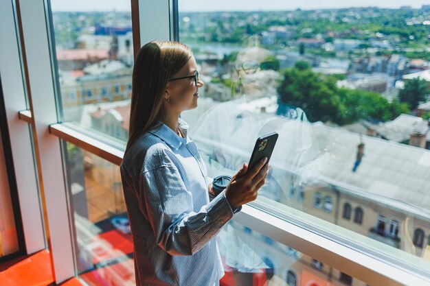 Blogueira feminina atraente com um smartphone moderno nas mãos fica na janela olhar pensativo e sonhar acordado segurando um telefone celular pondera uma ideia para uma publicação em redes sociais