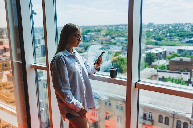 Blogueira feminina atraente com um smartphone moderno nas mãos fica na janela olhar pensativo e sonhar acordado segurando um telefone celular pondera uma ideia para uma publicação em redes sociais