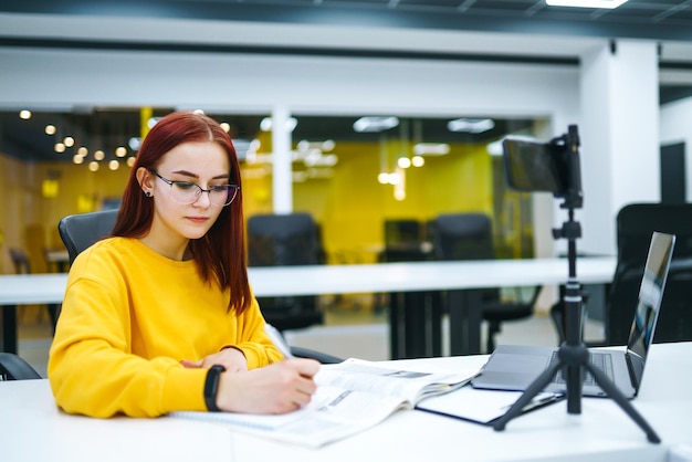 Blogueira feminina com laptop gravando vídeo enquanto está sentada no escritório, falando sobre negócios de inicialização.