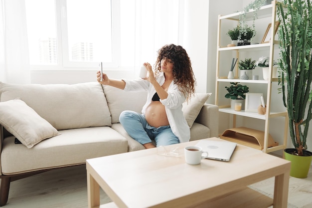 Blogueira grávida com um frasco de pílulas e vitaminas senta-se no sofá em casa e tira fotos