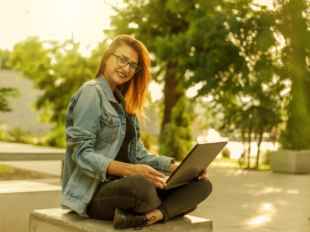 Blogueira jovem moderna em uma jaqueta jeans e óculos tem um laptop nas mãos no parque. Aprendizagem online, blogs