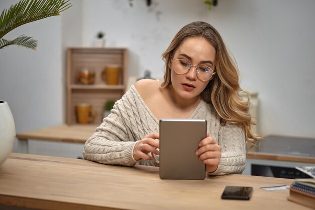 Blogueira loira de óculos camisola de malha bege segurando um tablet sentado na cozinha na mesa de madeira com livro cadernos smartphone cartão plástico e palmeira em pote branco nele closeup