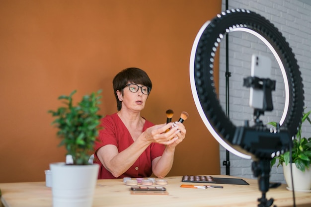 Blogueira ou influenciadora de beleza feminina de meia-idade com vídeo de gravação de produtos de maquiagem no quarto em casa