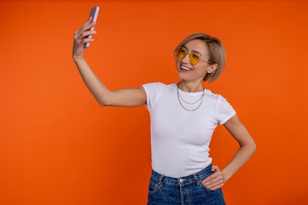 Foto blogueira satisfeita com camiseta branca e jeans fazendo selfie em smartphone isolada sobre fundo laranja