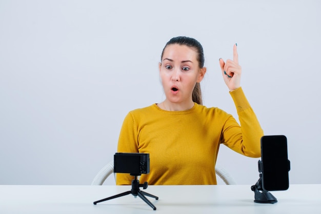 Blogueira surpresa está apontando para cima com o dedo indicador sentando-se na frente de sua mini câmera em fundo branco