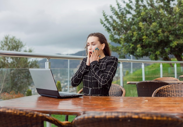 Blogueira surpresa está segurando as mãos nas bochechas olhando para a tela do laptop no fundo da visão da natureza