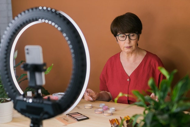 Blogueiro de beleza feminina de meia-idade ou influenciador com produto de maquiagem gravando vídeo no quarto em casa usando lâmpada de anel e smartphone