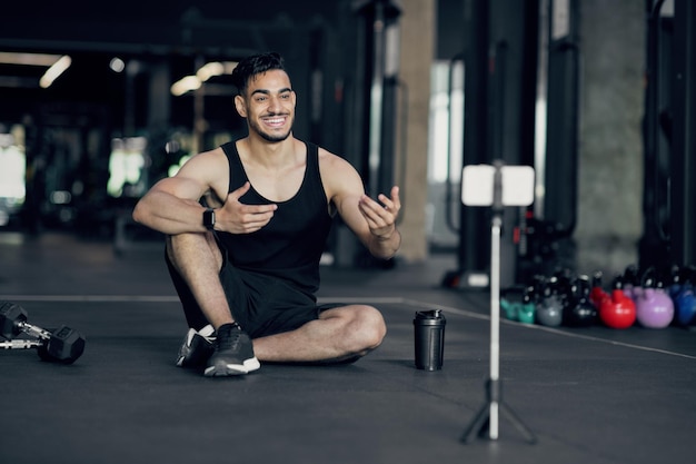 Blogueiro de fitness masculino árabe bonito gravando vídeo na câmera do smartphone no ginásio