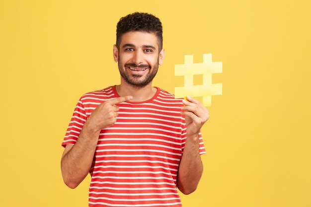 Blogueiro de homem barbudo positivo em camiseta listrada, apontando para o símbolo de hashtag amarelo em sua mão, recomendando postagens olhando para a câmera com sorriso dentuço Tiro de estúdio interno isolado em fundo amarelo