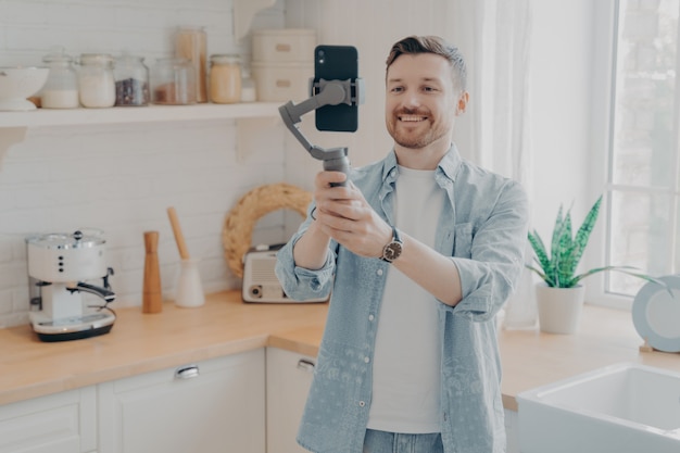 Blogueiro de jovem gravando videoblog ou transmissão ao vivo com smartphone e gimbal dentro de casa em pé no fundo da cozinha. vlogger de mídia social gravando vlog, streaming de podcast online no celular