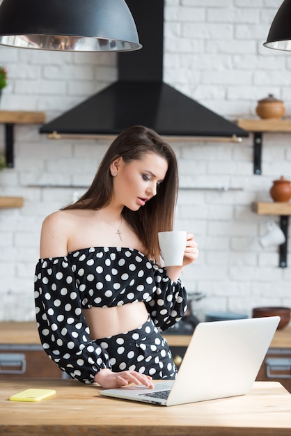 Blogueiro de menina morena atraente em um vestido de bolinhas, sentado na cozinha aconchegante, bebendo chá café e lendo notícias no laptop na mesa de madeira.