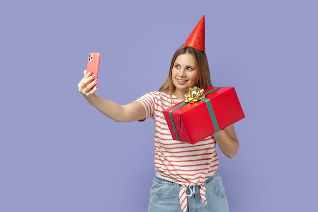 Blogueiro de mulher em cone de festa segurando caixa de presente e fazendo selfie ou fazendo videochamada