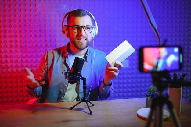 Blogueiro de tecnologia masculino gravando blog de vídeo ou revisão de vlog do novo smartphone mostrando-o na câmera no estúdio doméstico Conceito de trabalho de blog em casa Foco seletivo Web Banner