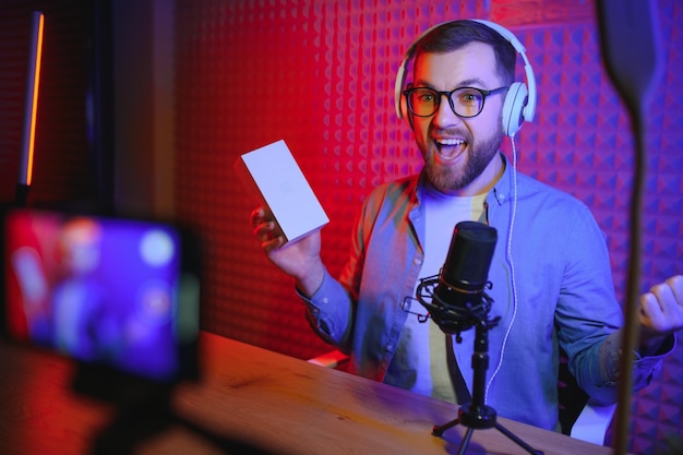 Blogueiro de tecnologia masculino gravando blog de vídeo ou revisão de vlog do novo smartphone mostrando-o na câmera no estúdio doméstico Conceito de trabalho de blog em casa Foco seletivo Web Banner