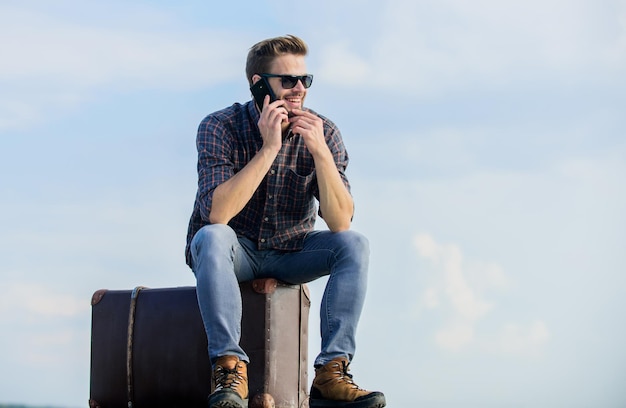 Blogueiro de viagens Homem sentado na mala antes da viagem Instalações de transporte Viajante bonito Cara ao ar livre com mala vintage Conceito de bagagem Chamando táxi Agência de viagens Viagem com bagagem