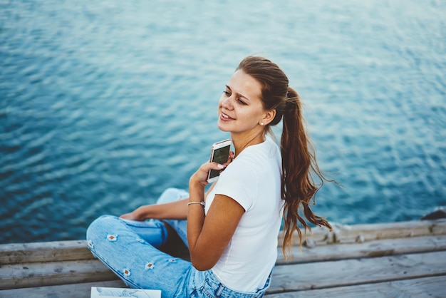 blogueiro sorridente olhando de lado sentado no cais de madeira e criando publicação para rede social