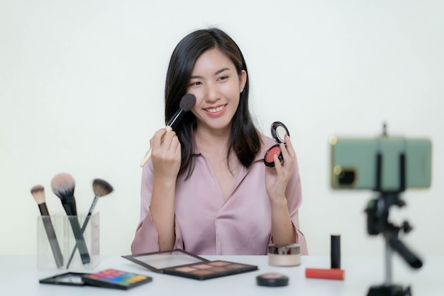 Una bloguera asiática bloguera de belleza grabando un video de ella con un lápiz labial naranja en un estudio.