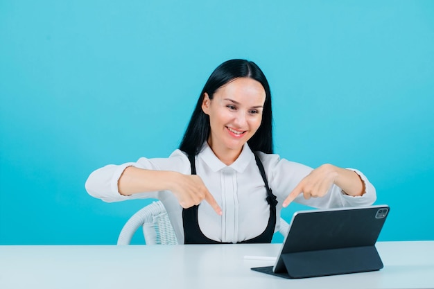 La bloguera sonriente está mirando la tableta apuntando hacia abajo con los dedos índices sobre fondo azul.