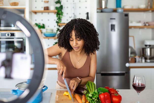 Bloguero de comida alegre usando delantal de pie en la mesa en la cocina moderna cortando verduras filmando este proceso en la cámara