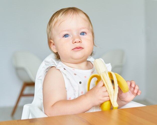 Blond blauäugiges Kleinkind hält und beißt Bananen gesunder Snack für die Kleinen